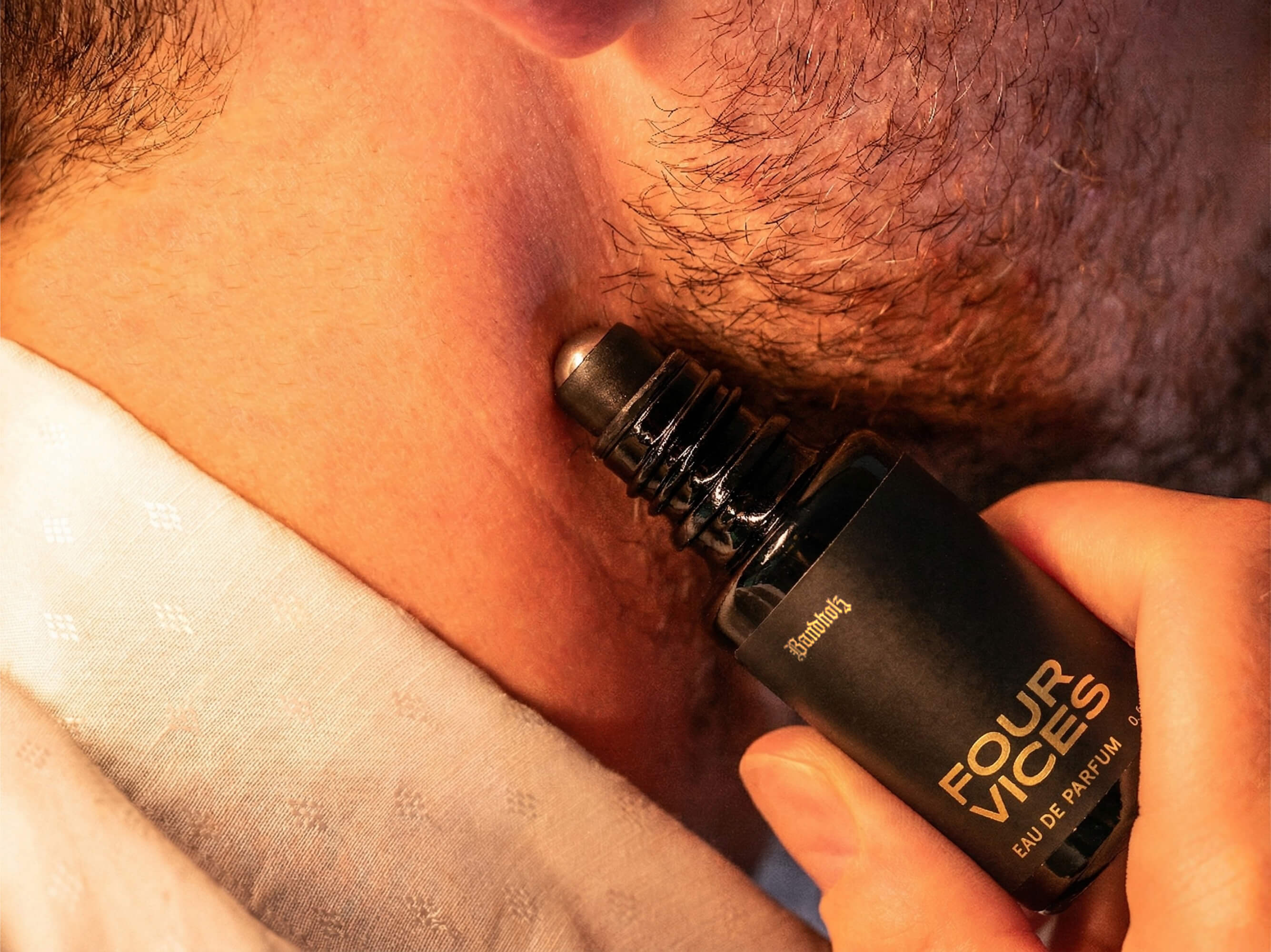 Close-up of stubbled lower face with Bandholz Four Vices Eau de Parfum roll-on bottle held against the skin.