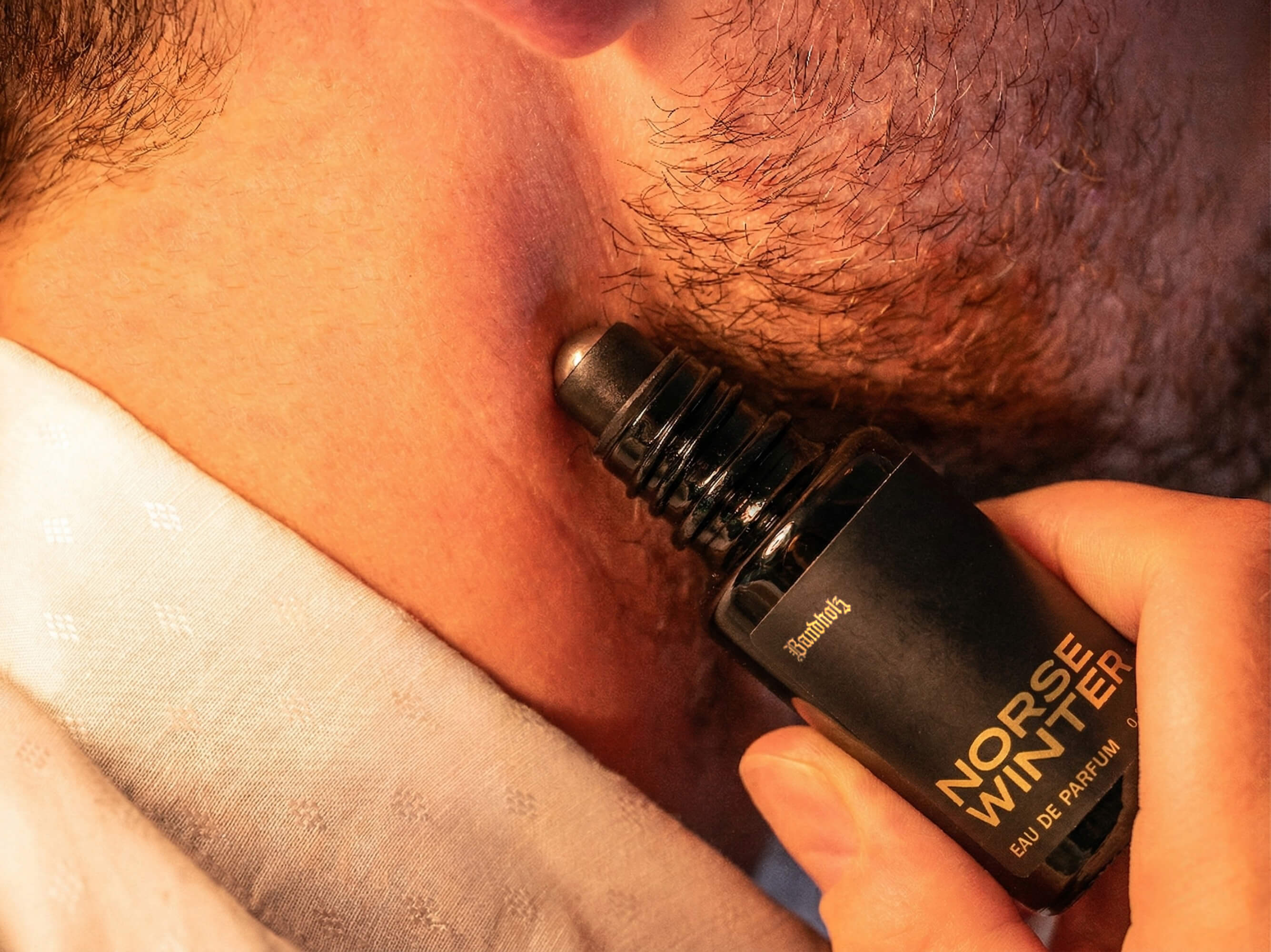 Close-up of stubbled lower face with Bandholz Norse Winter Eau de Parfum roll-on bottle held against the skin.