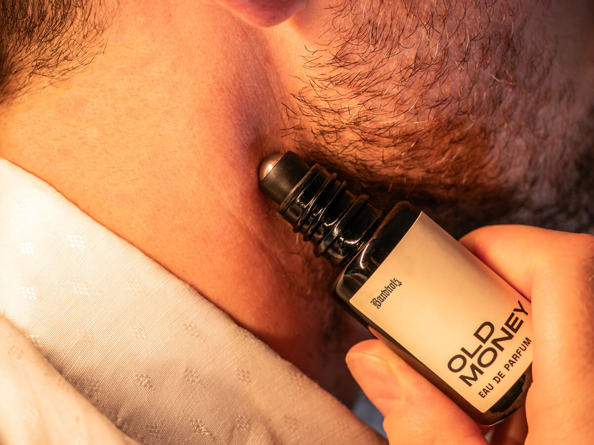 Close-up of stubbled lower face with Bandholz Old Money Eau de Parfum roll-on bottle held against the skin.