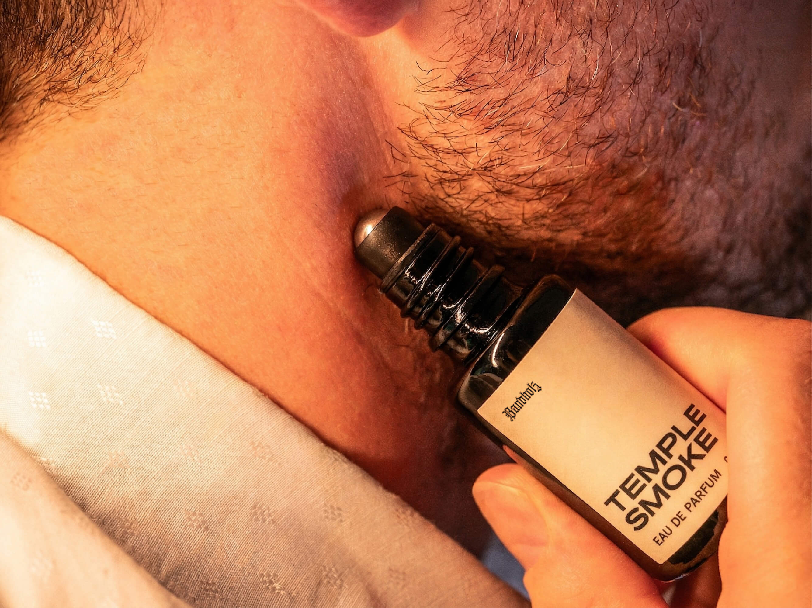 Close-up of stubbled lower face with Bandholz Temple Smoke Eau de Parfum roll-on bottle held against the skin.