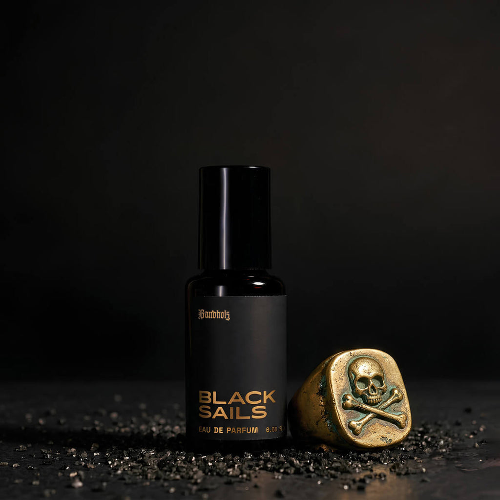 Black Sails Eau de Parfum bottle by Bandholz on a dark surface with scattered black grains and a gold skull-and-crossbones ring beside it. Image suggests a bold maritime fragrance with notes of tobacco, bourbon, spice, and ambergris.